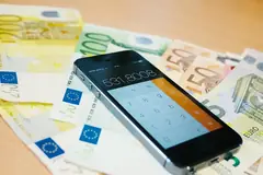 Смартфон с отворен калкулатор върху разпилени евро банкноти от 5, 50, 100 и 200 евро на дървена маса