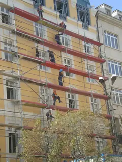Строители с каски и работни униформи на скеле пред жълта фасада на стара многоетажна сграда